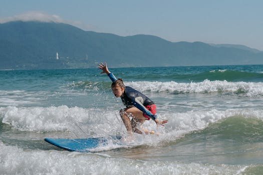 A woman riding a surfboard on ocean waves against a mountainous coastline backdrop.
